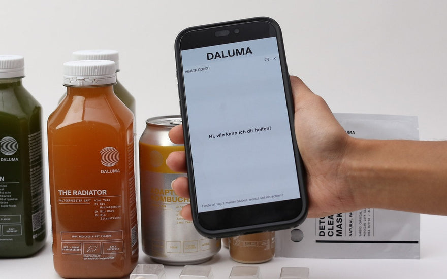 Smartphone mit dem DALUMA Health Coach Chat vor kaltgepressten Säften, Kombucha und Detox Clearing Mask.