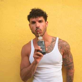 Ein Mann mit lockigem Haar und einem tätowierten Ärmel auf dem linken Arm steht vor einer gelben Wand, trägt ein weißes Tank-Top und trinkt ein dunkles Getränk aus einer Glasflasche durch einen Strohhalm.