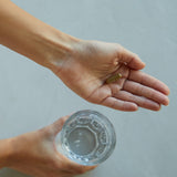 Eine Person hält ein Glas Wasser in der einen Hand und eine einzelne Kapsel der DALUMA LONGEVITY SUPPLEMENT PROTOKOLL in der anderen, vor einem schlichten hellen Hintergrund, der einen einfachen Schritt in ihrer Wellness-Routine illustriert.