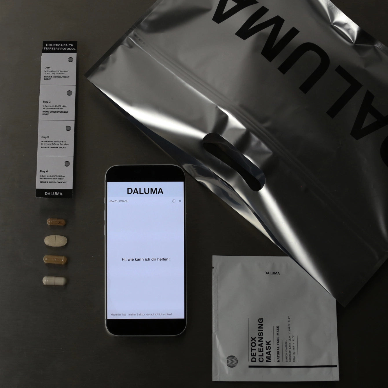 Auf einer flachen Unterlage befinden sich vier Ergänzungskapseln, eine Beipackzettel, ein Smartphone mit DALUMA, eine DALUMA Metalltasche und eine DETOX CLEANSING MASK - alles, was Sie für Ihre BESSERE SAFTKUR PLUS - 3 TAGE by DALUMA benötigen.