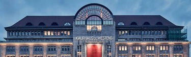 Ein großes, historisches Gebäude mit dem Kaufhaus des Westens an der Fassade, Rundbogenfenstern und einer zentralen Glaskuppel über einem roten Eingang, der abends beleuchtet wird.