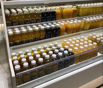 Reihen von abgefüllten Säften in einer Kühlvitrine, geordnet nach Farbe und Art. Die meisten Flaschen haben weiße Verschlüsse und enthalten orangefarbene, gelbe oder dunkelfarbige Flüssigkeiten.