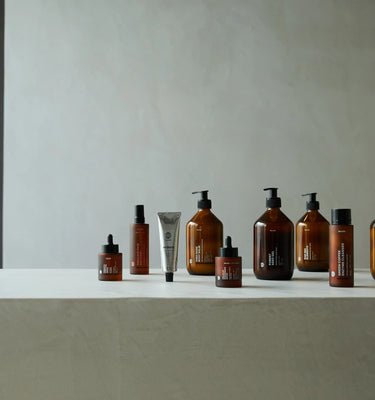 Ein minimalistisches Display aus bernsteinfarbenen Flaschen und Gläsern mit schwarzen Pumpen und Deckeln sowie einem Metallrohr, die auf einer hellen Fläche vor einem schlichten, neutralen Hintergrund angeordnet sind.