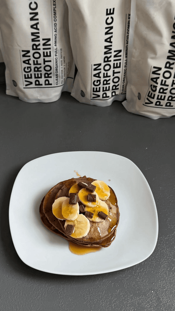 Ein weißer Teller mit Pfannkuchen, die mit Bananenscheiben, Schokoladenstückchen und Sirup belegt sind, steht auf einer dunklen Fläche. Dahinter stehen aufrecht stehende Tüten mit der Aufschrift Vegan Performance Protein.