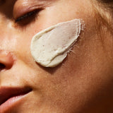 Nahaufnahme des Gesichts einer Person mit geschlossenen Augen, auf deren Wange ein dicker Klecks dailumade.comEP HYDRATION FEUCHTIGKEITSMASKE mit natürlichen Zutaten und Shea Butter aufgetragen ist; die glatte Haut leuchtet im Sonnenlicht.