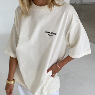 Eine Person trägt das DALUMA JOSEPH ATELIER X DALUMA SELFCARE SUNDAY TEE, ein nachhaltiges cremefarbenes T-Shirt aus Bio-Baumwolle in Übergröße mit Brustaufdruck, gepaart mit einer weißen Hose, während sie in einem Haus steht und eine Hand in die Hüfte stemmt.