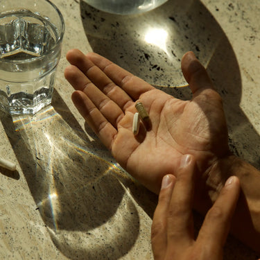 Eine Person hält drei dailumade.comFENCE DUO Kapseln in ihrer offenen Handfläche neben einem Glas Wasser auf einer sonnenbeschienenen, gesprenkelten Arbeitsplatte. Das Licht des Glases reflektiert auf der Oberfläche und symbolisiert die tägliche Unterstützung des Immunsystems.