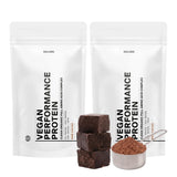 Zwei weiße DALUMA VEGANES PERFORMANCE PROTEIN KAKAO DOUBLE-Beutel stehen hinter Schokoladenwürfeln und einem Metalllöffel mit Bio-Proteinpulver, der den reichen Rohkakaogeschmack dieses veganen Proteins zur Geltung bringt.