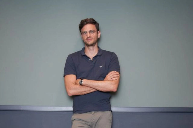 Ein Mann mit kurzen braunen Haaren und Brille steht mit verschränkten Armen vor einer schlichten graugrünen Wand und trägt ein dunkles Poloshirt und eine khakifarbene Hose.