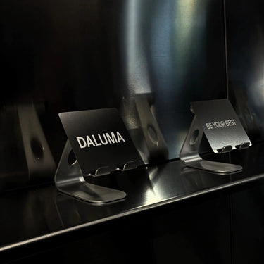 Zwei Metallständer mit den Aufschriften DALUMA und BE YOUR BEST stehen auf einer glänzenden schwarzen Oberfläche vor einer reflektierenden Wand - ideal für die Präsentation Ihres DALUMA THE GOOD VOUCHER GUTSCHEIN 30€, 50€ oder 100€.