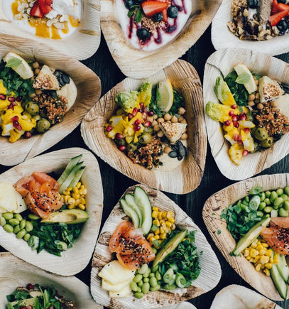 Eine Draufsicht auf mehrere dreieckige Holzteller, die mit bunten Speisen gefüllt sind, darunter Salate, Gemüse, Körner, Grünzeug, Avocado, Mais, Kichererbsen, Grünzeug, Lachs, Samen und Joghurt mit Obst.