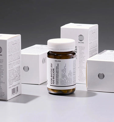 Eine braune Flasche mit der Aufschrift SYM-BIOTIC 20/100 BILLION ist umgeben von vier weißen Produktschachteln mit demselben Branding, die auf einer grauen Fläche unter heller Beleuchtung angeordnet sind.