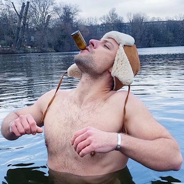 Ein Mann steht brusttief in einem See, lehnt sich zurück und hält eine kleine Flasche DALUMA BURNING MAN INGWERSHOT 24 PACK in den Mund. Im Hintergrund sind blattlose Bäume und ein wolkenverhangener Himmel zu sehen.