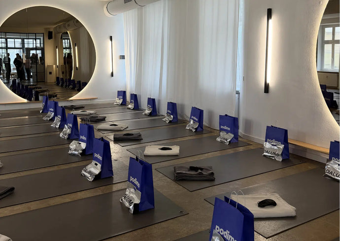 Ein modernes Yogastudio mit Reihen von Yogamatten, jede mit einer blauen Geschenktasche, einem Handtuch und einer gefalteten Decke. Der Raum verfügt über einen großen runden Spiegel, Bänke und sanfte Beleuchtung an weißen, geschwungenen Wänden.