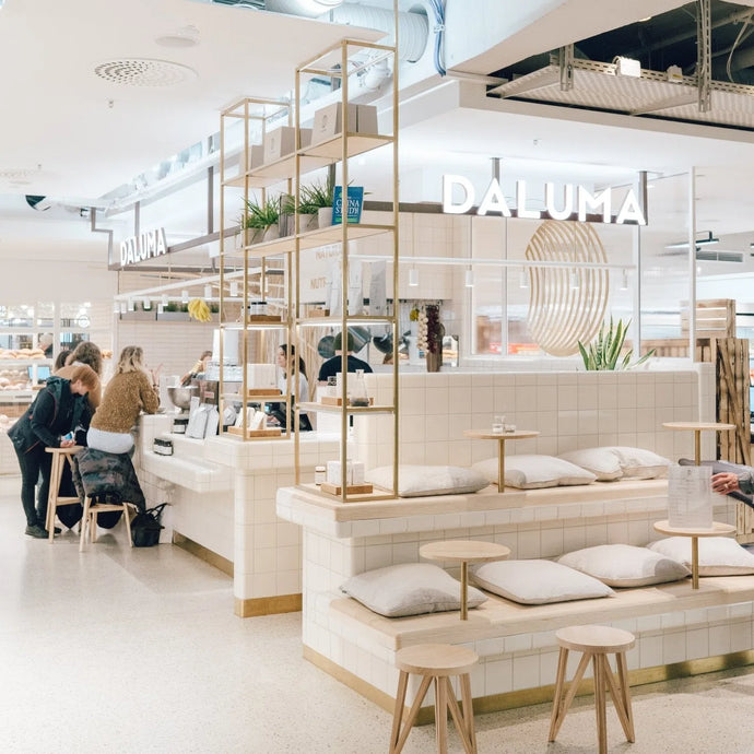 Ein modernes, minimalistisches Café namens DALUMA mit weißen Fliesen, hellen Holzmöbeln, kleinen runden Tischen und Sitzkissen. Mehrere Leute bestellen und sitzen. Der Raum wirkt hell und luftig.