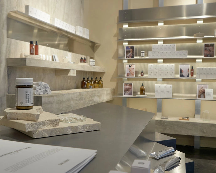 Ein modernes, minimalistisches Ladeninnere mit Steinregalen, in denen Hautpflege- und Wellnessprodukte in Flaschen und Schachteln ausgestellt sind, sanft beleuchtet vor beigen Wänden und Beton. Im Vordergrund befindet sich eine Arbeitsplatte mit Produktproben.