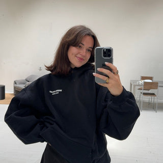 Eine junge Frau mit schulterlangen braunen Haaren macht ein Spiegel-Selfie in einem hellen, minimalistischen Raum. Sie trägt einen schwarzen Kapuzenpullover und lächelt, im Hintergrund sind ein Sofa und Stühle zu sehen.