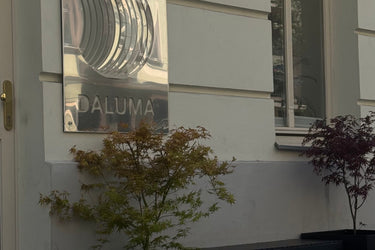 Ein glänzendes Metallschild mit der Aufschrift DALUMA ist an einer hellen Hauswand in der Nähe eines Fensters angebracht. Vor dem Schild stehen zwei getopfte Ahornpflanzen mit grünen und roten Blättern.