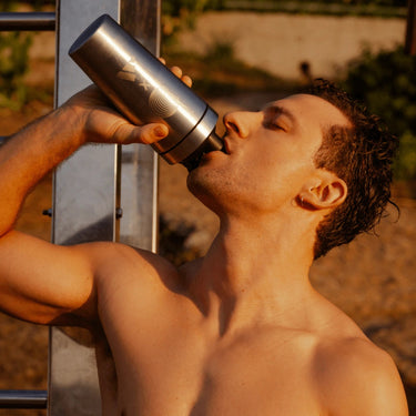 Ein hemdsärmeliger Mann trinkt im Freien aus einer silbernen Flasche, das Sonnenlicht beleuchtet ihn, während er sich an Metallstangen lehnt - gestärkt nach einem Training mit DALUMA VEGANES PERFORMANCE PROTEIN ALL FLAVORS.