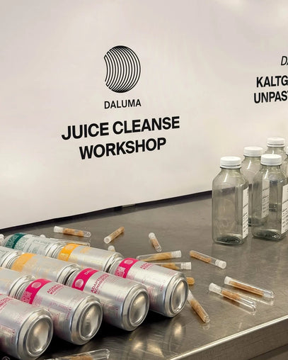 Mehrere Dosen, kleine Fläschchen mit Flüssigkeitsproben und Glasflaschen sind auf einem Tresen angeordnet. Auf einem Schild im Hintergrund steht DALUMA JUICE CLEANSE WORKSHOP.