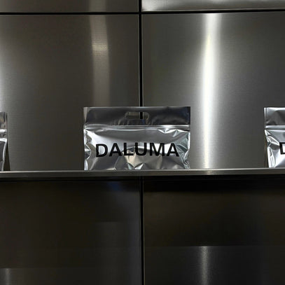 Ein metallisches Regal mit einer silbern glänzenden Tasche, auf der in fetten schwarzen Buchstaben DALUMA steht, vor einem Hintergrund aus rostfreiem Stahl. Zwei ähnliche Tüten sind teilweise auf beiden Seiten sichtbar.