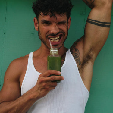 Ein Mann in einem weißen Tank Top mit lockigem Haar und Tattoos zwinkert und lächelt verspielt, hält eine Flasche DALUMA GRÜNE SAFTKUR PLUS - 5 TAGE mit einem Strohhalm in der Hand, den Arm über den Kopf erhoben, und steht vor einer grünen Wand.