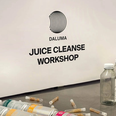 Ein Schild mit der Aufschrift DALUMA JUICE CLEANSE WORKSHOP prangt an einer weißen Wand. Davor liegen leere Flaschen, Dosen und kleine Fläschchen auf einem Tisch verstreut.
