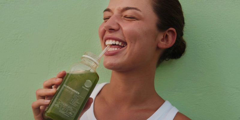 Eine lächelnde Frau in einem weißen Tank-Top trinkt grünen Saft aus einer Glasflasche mit einem Strohhalm und steht vor einer hellgrünen Wand.