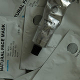 Mehrere DALUMA HYDRATING ESSENTIALS Produkte, darunter eine Metalltube und flache Päckchen mit der Aufschrift Natural Face Mask und Inhaltsstofflisten, sind auf einer wassergetränkten Fläche verstreut. Das DALUMA-Logo erscheint auf jedem Produkt.