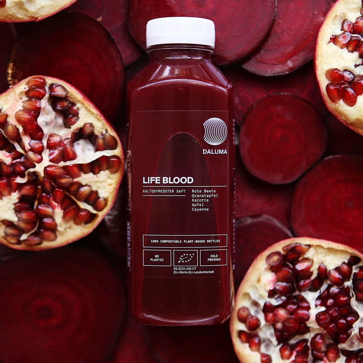 Eine Packung DALUMA LIFE BLOOD POWER SAFT 6 PACK, mit Kaltgepresster Saft, ist umgeben von Granatapfel- und Rübenschnitzen. Das Etikett hebt Zutaten wie rote Beete Saft und Granatapfelsaft hervor und betont die umweltfreundliche Verpackung.