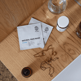 Drei DALUMA CLEANSE AND HYDRATE MASK DUO-Sachets werden zusammen mit einem kleinen Tiegel, einem klaren Glas, einem braunen Behälter und Goldschmuck auf einer Holzfläche neben einer weißen Arbeitsplatte präsentiert.