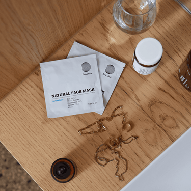 Drei DALUMA CLEANSE AND HYDRATE MASK DUO-Sachets werden zusammen mit einem kleinen Tiegel, einem klaren Glas, einem braunen Behälter und Goldschmuck auf einer Holzfläche neben einer weißen Arbeitsplatte präsentiert.