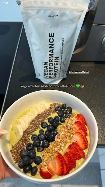 Eine Hand hält eine Smoothie-Schale mit Bananenscheiben, Leinsamen, Blaubeeren, Granola und Erdbeerscheiben. Dahinter liegt eine Tüte Daluma Vegan Performance Protein auf einem Küchentisch.