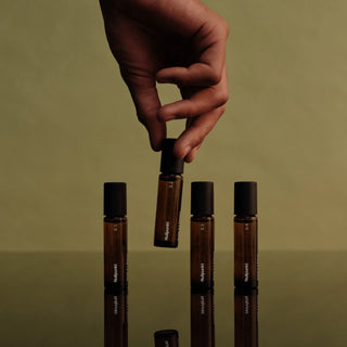 Eine Hand hält eine von vier kleinen braunen Glasflaschen mit schwarzen Verschlüssen, die auf einer spiegelnden Fläche vor einem schlichten olivgrünen Hintergrund aufrecht stehen.