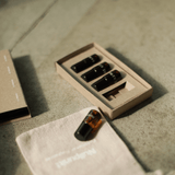 Vier kleine schwarze Fläschchen aus dem Nullpunkt Discovery Set von Nullpunkt sind auf einer strukturierten Oberfläche in einer Pappschachtel angeordnet. Eine Flasche steht außerhalb der Schachtel neben einem hellen Stoffbeutel mit schwacher Aufschrift.