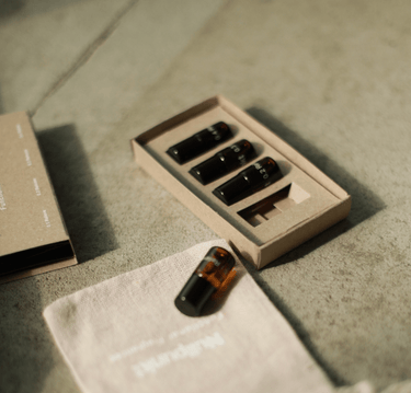 Vier kleine schwarze Fläschchen aus dem Nullpunkt Discovery Set von Nullpunkt sind auf einer strukturierten Oberfläche in einer Pappschachtel angeordnet. Eine Flasche steht außerhalb der Schachtel neben einem hellen Stoffbeutel mit schwacher Aufschrift.