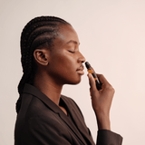 Eine Frau mit geflochtenem Haar und geschlossenen Augen hält einen Nullpunkt GRADE 0.1 RELIEVE Aromatherapie-Ölroller an ihre Nase, als ob sie daran riechen würde, während sie einen schwarzen Blazer vor einem hellen Hintergrund trägt.