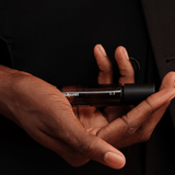 Eine Person in einem dunklen Anzug hält einen braunen Glasflakon des Parfums Nullpunkt GRADE 0.3 REFOCUS in der Hand, wobei ihre Finger den Duft von Nullpunkt vor einem schattigen Hintergrund sanft umschmeicheln.