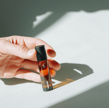 Eine Hand hält einen Flakon NULLPUNKT GRADE 0.3 REFOCUS von Nullpunkt aus Braunglas mit schwarzem Verschluss, der im hellen Sonnenlicht einen scharfen Schatten auf eine weiße Oberfläche wirft.