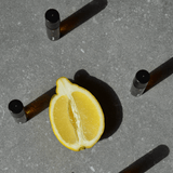 Eine halbierte Zitrone auf einer grauen Fläche ist von vier braunen Nullpunkt NULLPUNKT GRADE 0.4 REFUEL-Flaschen mit schwarzen Verschlüssen umgeben, die jeweils lange Schatten werfen.