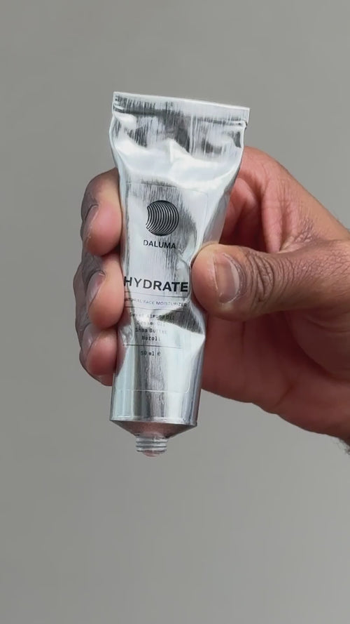 Eine Hand hält eine Metalltube mit der Aufschrift DALUMA HYDRATE vor einem schlichten, hellen Hintergrund. Die Tube scheint für ein Hautpflege- oder Kosmetikprodukt zu sein.