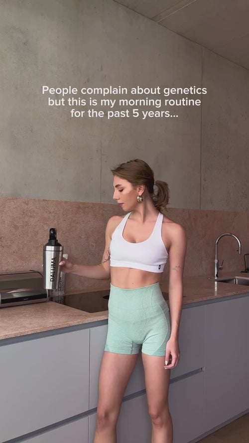 Eine Frau in Sportkleidung steht in einer modernen Küche und hält eine Mixerflasche. Der Text über ihr lautet: "Die Leute beschweren sich über Genetik, aber das ist meine Morgenroutine der letzten 5 Jahre...