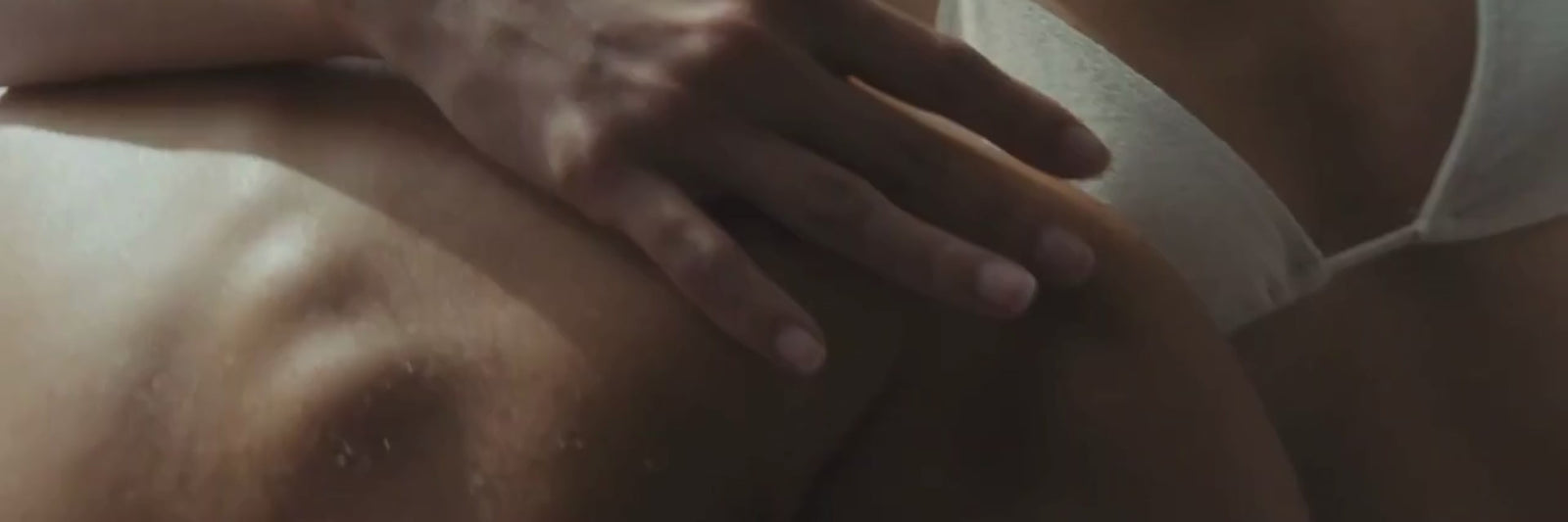 Eine Nahaufnahme der Hand einer Person, die auf dem nackten Rücken einer anderen Person ruht; die zweite Person trägt einen weißen BH, und die Szene ist sanft beleuchtet, was die Struktur der Haut und die sanfte Berührung betont.