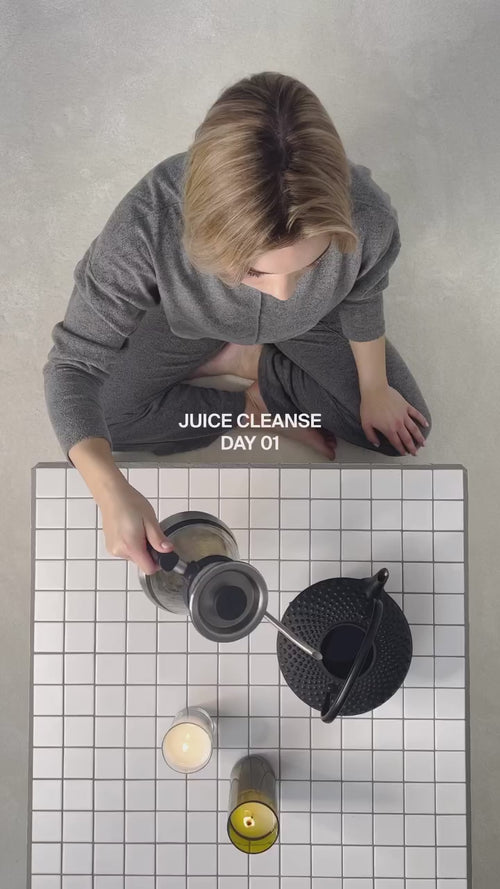 Eine Person in grauer Loungewear sitzt auf dem Boden neben einem gekachelten Tisch und gießt Flüssigkeit aus einer schwarzen Teekanne ein. Auf dem Tisch stehen Kerzen. Der Text lautet: "JUICE CLEANSE DAY 01.".