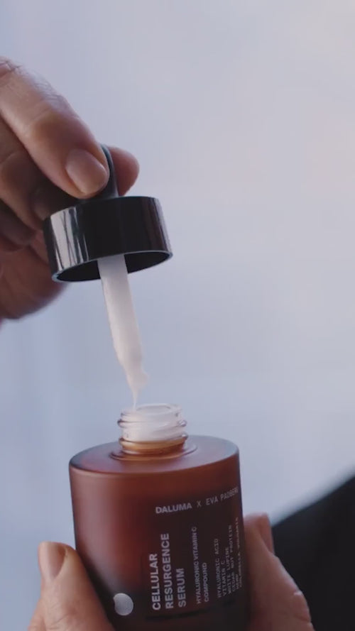 Eine Hand hält eine braune Flasche mit einem Hautpflegeserum, während eine andere Hand eine Pipette mit weißem Serum aus der Flasche hebt, vor einem weichen, hellen Hintergrund. Auf dem Etikett steht "Cellular Resurgence Serum".