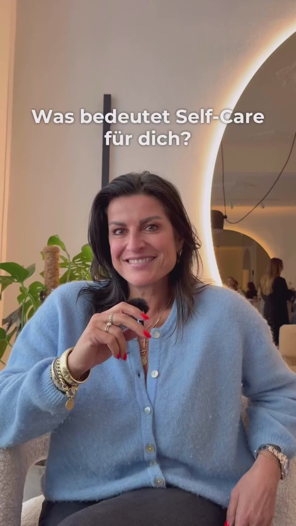 Eine lächelnde Frau in einer blauen Strickjacke sitzt mit einem Mikrofon in der Hand. Der Text über ihr lautet: Was bedeutet Self-Care für dich? Im Hintergrund sind Pflanzen und eine sanft beleuchtete moderne Einrichtung zu sehen.