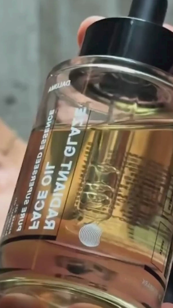Eine Hand hält eine Klarglasflasche mit einer bernsteinfarbenen Flüssigkeit und einem Tropfer mit der Aufschrift GRAVITY FACE OIL in fetten Buchstaben in der Nähe des Gesichts einer Person im verschwommenen Hintergrund.