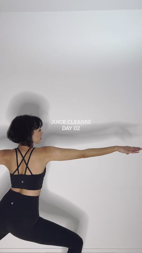 Eine Frau in schwarzer Trainingskleidung macht eine Yoga-Pose vor einer weißen Wand. Der Text an der Wand lautet JUICE CLEANSE DAY 02.