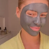 Eine Frau mit einer grauen Gesichtsmaske und einem gelben Bademantel lächelt sanft. In einem Regal im Hintergrund stehen Hautpflegeprodukte.