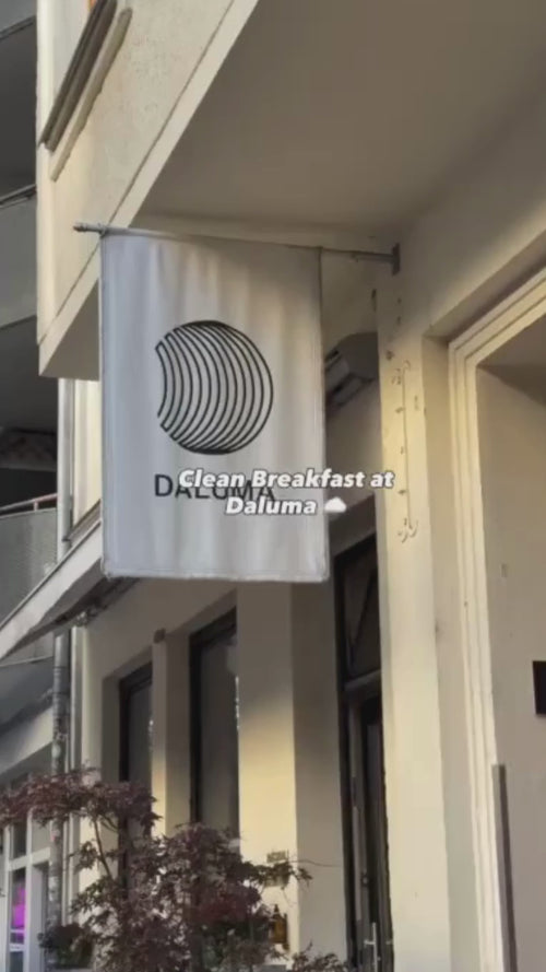 Eine weiße Flagge mit einem runden schwarzen Logo und dem Wort DALUMA hängt vor einem Gebäude. Der überlagerte Text lautet: "Sauberes Frühstück bei Daluma". Einige Pflanzen sind in der Nähe des Eingangs zu sehen.